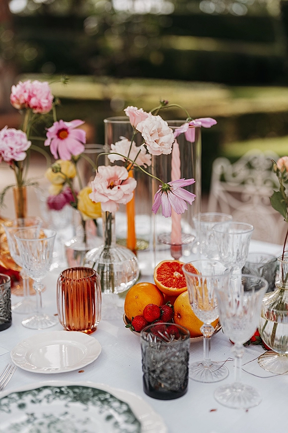 Reception tablescape with an outdoor reception table featuring wildflower bud vases, pink tapers, fruit centerpiece, and glassware on a garden lawn