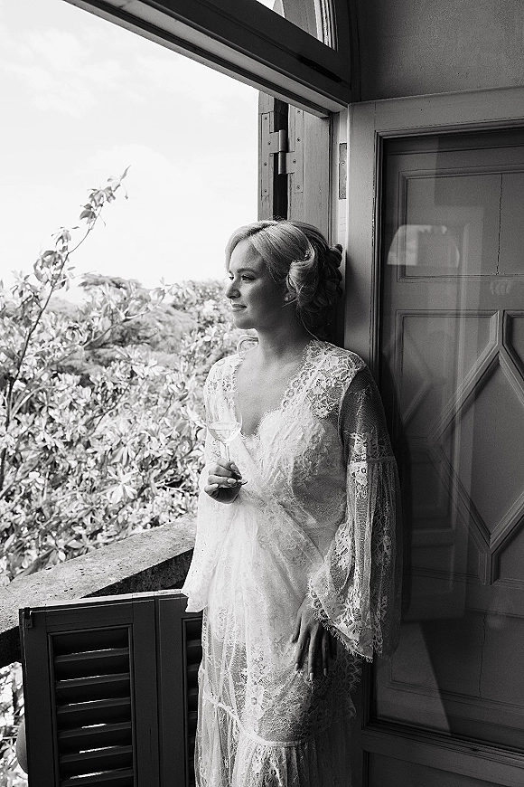 Bridal portrait of a bride looking out window in a lace wedding dress with long sleeves, holding a champagne flute by the balcony railing