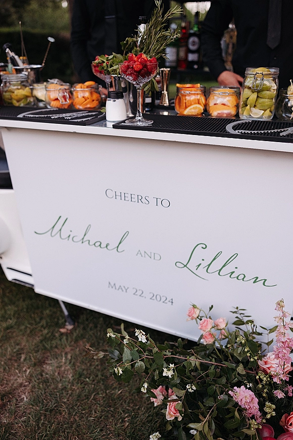 Wedding bar setup with a mobile wedding bar sign, glassware, liquor bottles, and garnish jars on an outdoor lawn with bartenders in black