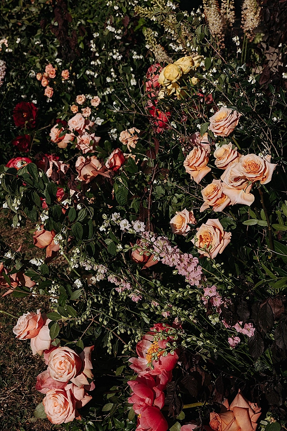 Wedding florals and a garden rose arrangement of peach roses, pink peonies, and greenery with lavender stock accents on grass in foliage