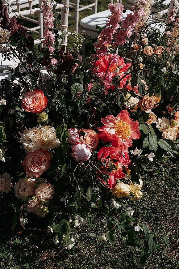 Ceremony aisle flowers in coral and pink roses and peonies with greenery garland lining the grass aisle beside white Chiavari chairs