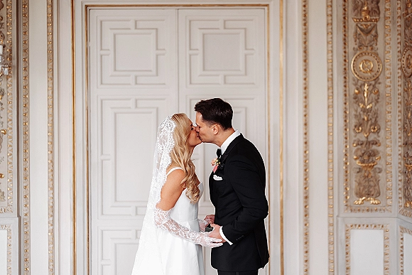 Wedding kiss portrait of bride and groom kissing in side profile, her lace veil framing a black tuxedo groom before ornate white doors