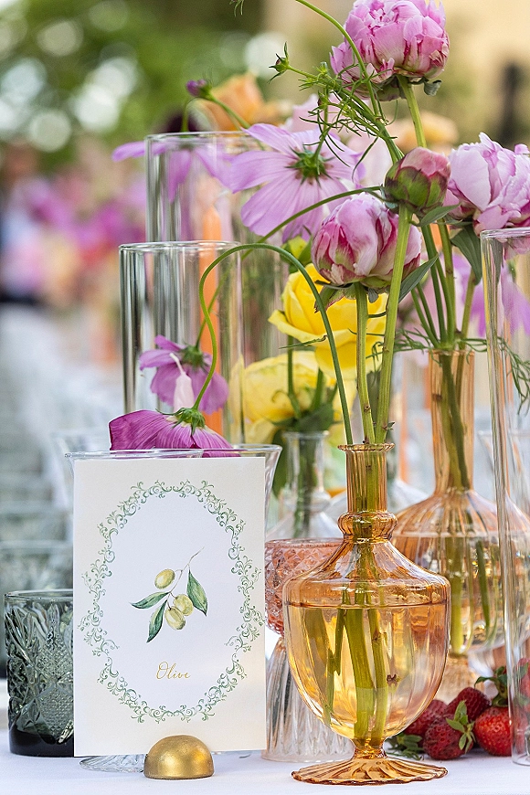 Reception tablescape with a wedding table centerpiece of pink peonies, cosmos and yellow roses in amber and clear vases, greenery behind