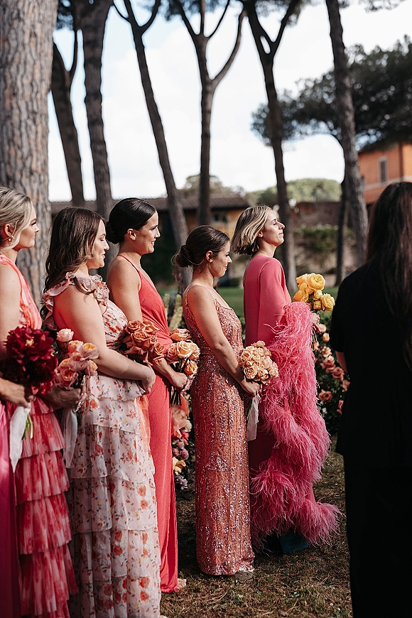 Wedding guests in ceremony guest attire hold rose bouquets, wearing floral and sequined dresses with a pink feather wrap on a garden lawn