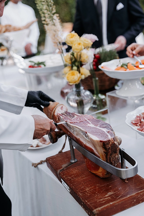 Wedding food station with a jamon carving station as gloved chef slices cured ham beside bread basket and floral centerpiece outdoors