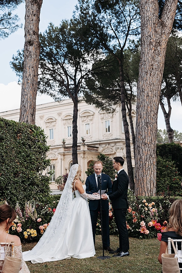 Wedding vows as bride in a long lace veil and groom in a black tux hold hands at an outdoor garden ceremony on a lawn with hedges and pines