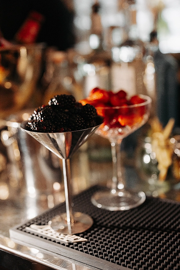 Wedding cocktail bar featuring signature wedding cocktails in a martini glass and coupe glass with blackberry and strawberry garnish on a metal counter