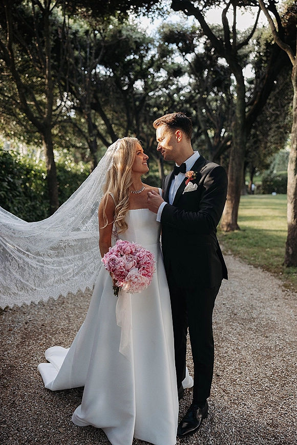 Couple portrait of bride in strapless gown and lace veil holding a pink bouquet, beside groom in tuxedo on a tree-lined park path