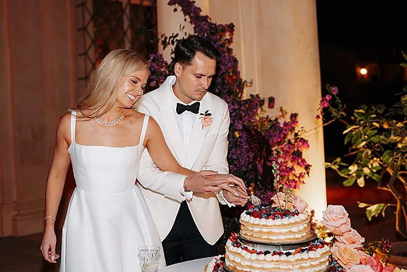 Wedding cake cutting as bride and groom slice a berry-topped naked cake on a stand under warm evening lights and columns, with roses
