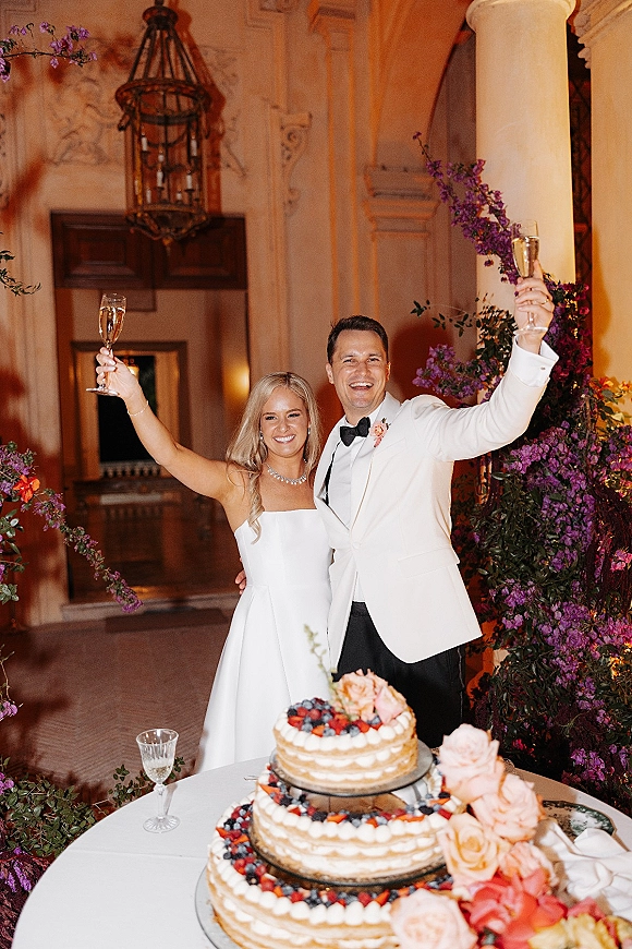 Wedding reception toast as bride in strapless gown and groom in white dinner jacket clink champagne flutes by berry-topped cake under chandelier