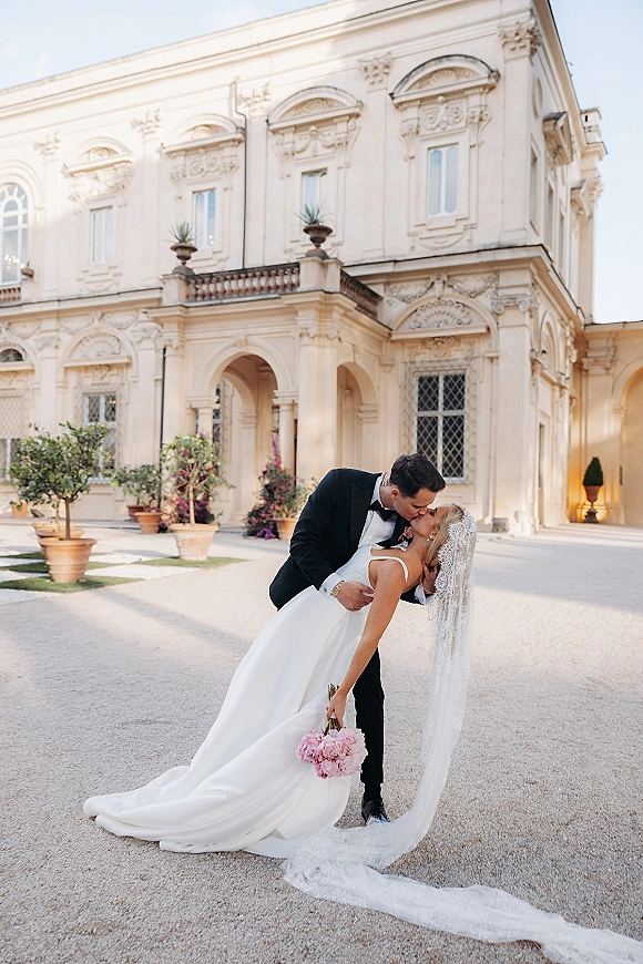 Wedding kiss portrait of groom dipping bride in a lace veil and train, holding a pink bouquet, in a historic courtyard archway