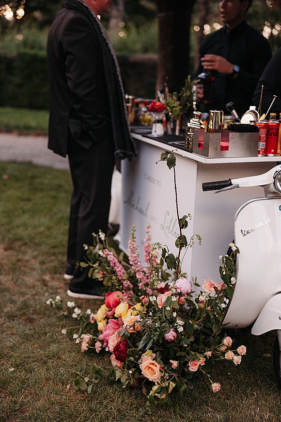 Wedding bar setup with a mobile wedding bar scooter cart, cocktail shakers and glassware, styled with floral arrangement on a garden lawn at dusk