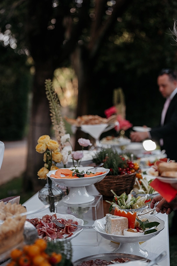 Wedding food display with a wedding charcuterie table of cured meats, cheese wheel, bread, fruit, and red napkins on a garden buffet table