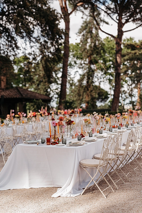 Reception tablescape with long banquet table wedding setup, white linens, place settings, amber bud vases, colorful flowers and taper candles in a garden pavilion
