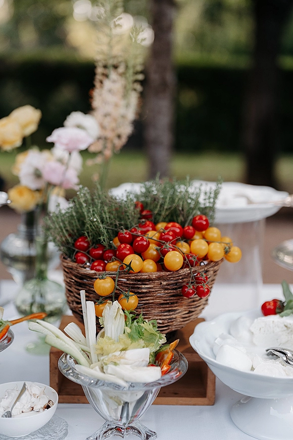 Wedding food table with wedding grazing table spread of crudites, dips, and a wicker basket of cherry tomatoes on a white cloth in a garden setting