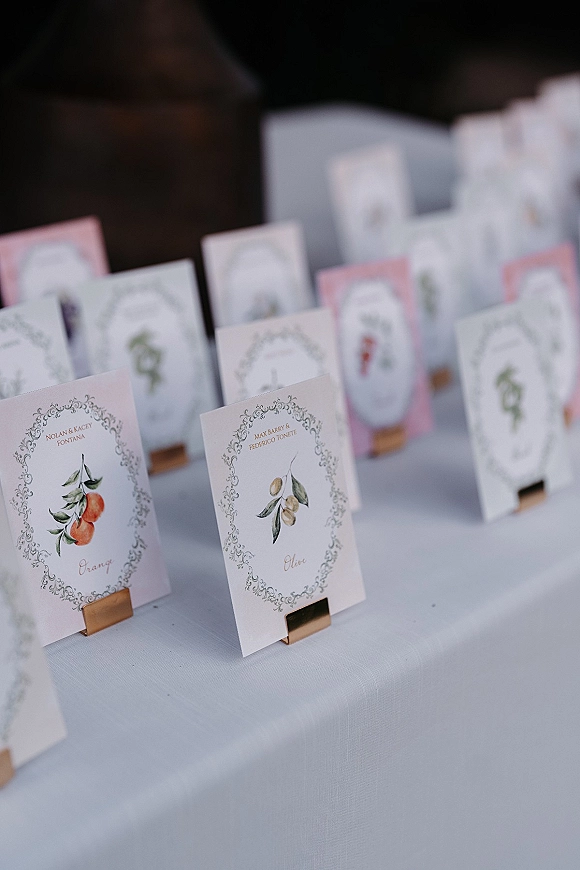 Wedding escort cards in an escort card display, printed with fruit illustrations and ornate borders in brass holders on a linen table