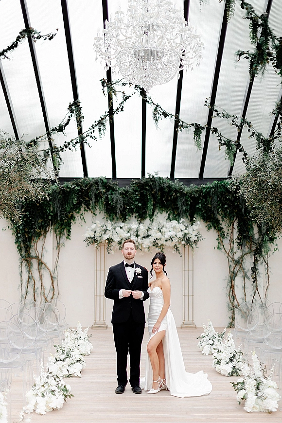 Bride and groom portrait with black tuxedo groom beside bride in a strapless slit gown under a crystal chandelier in a glass-roof venue