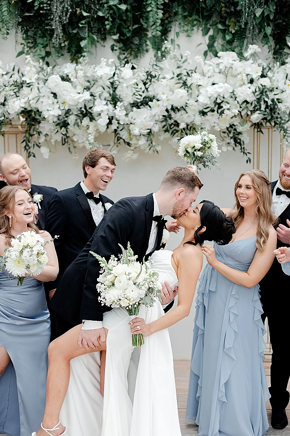 Wedding party photo of bride and groom kiss in a dipped pose, surrounded by bridesmaids in blue and tuxedoed groomsmen by a greenery wall