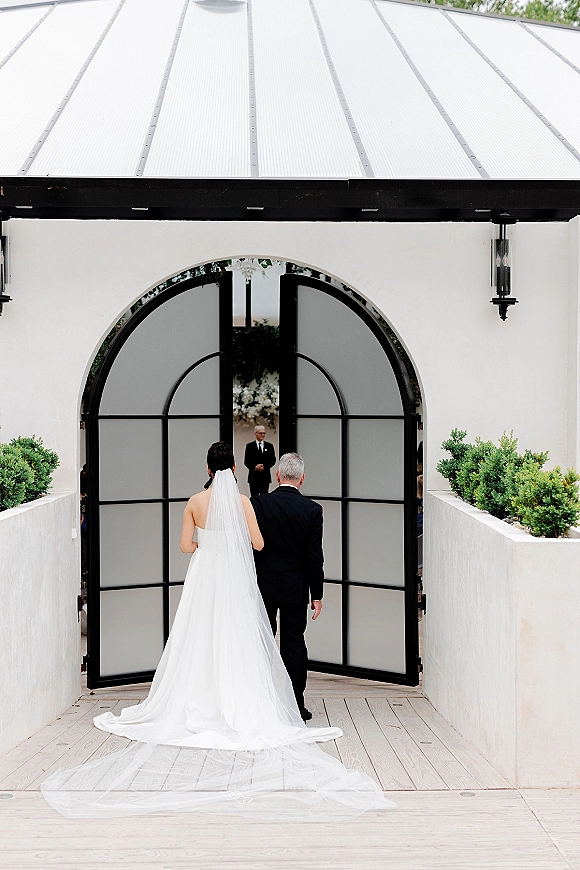 Wedding processional as bride walking down aisle from behind with her father, cathedral veil and long train at white stucco arched doors