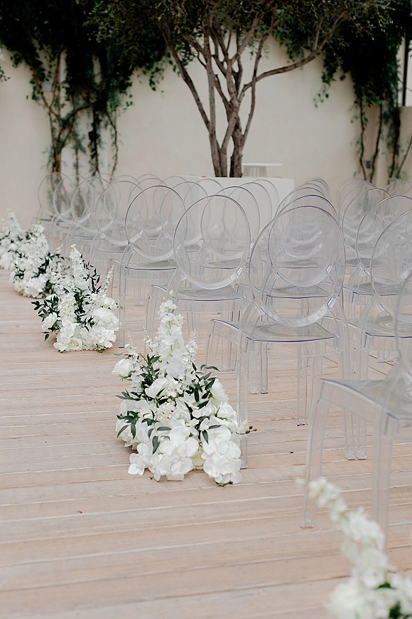 Ceremony aisle decor with white aisle flowers and greenery lining a wooden deck aisle beside clear acrylic chairs against a stucco wall