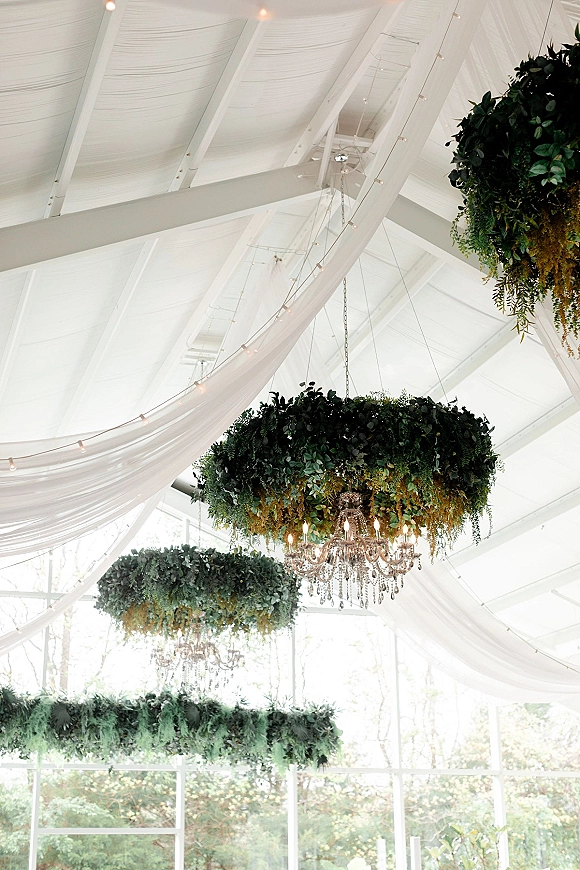 Hanging floral chandelier with greenery installation and crystal chandelier, framed by white vaulted beams, drapery, and string lights