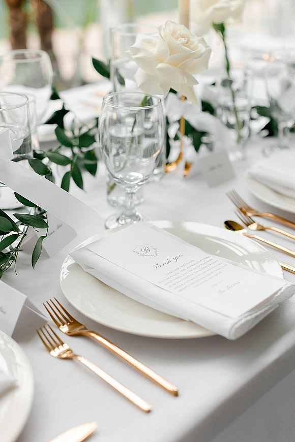 Reception tablescape with wedding place setting, white rose and greenery garland, place cards, thank you note, gold flatware and goblets
