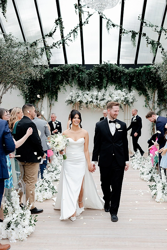 Wedding recessional as bride and groom walk down aisle holding hands, bouquet and rose petals under a glass ceiling with guests cheering