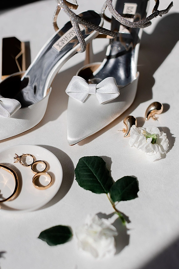 Bridal shoes in white satin wedding heels with bow and rhinestone straps, styled with rings and a white rose on a sunlit tabletop