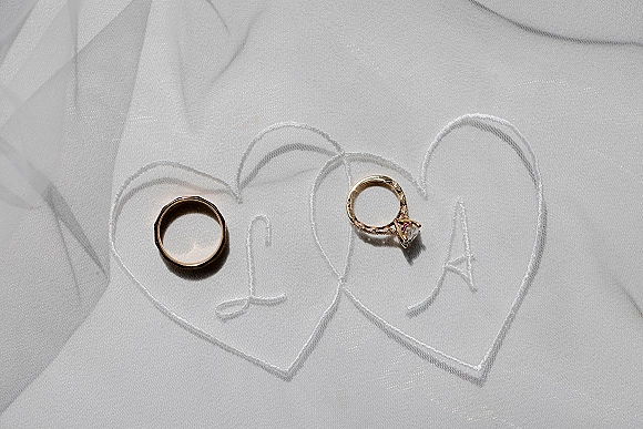 Wedding rings resting on a white veil, gold wedding band and diamond engagement ring framed by an embroidered heart outline in tulle