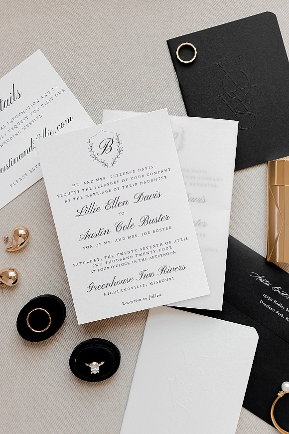 Wedding invitations arranged as a wedding invitation suite with black envelope, gold wax seal, rings, perfume and lipstick on linen backdrop