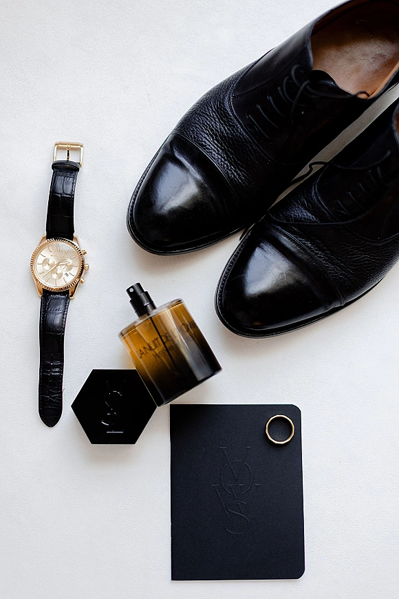 Groom details flatlay with black leather dress shoes, gold wristwatch, cologne bottle, and wedding ring on a white surface