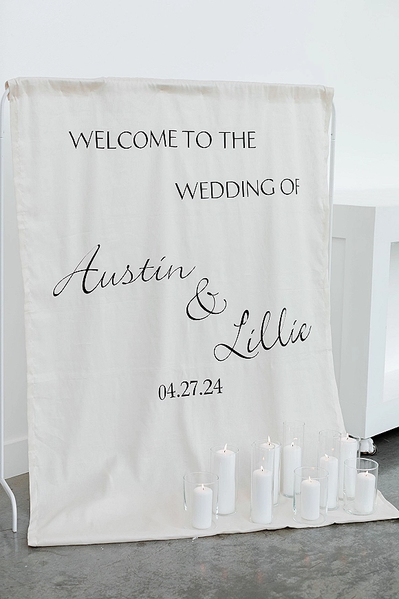 Wedding welcome sign with fabric backdrop and black calligraphy, set on a metal stand with white pillar candles and glass vases on concrete floor