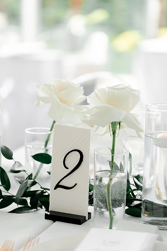 Wedding table number card in a black holder on a white tablecloth, with white roses, bud vases, greenery garland, and water glasses near windows