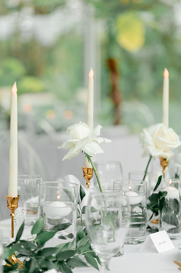 Reception tablescape with a wedding table centerpiece of white roses and greenery garland, plus gold candlesticks and taper candles by window light