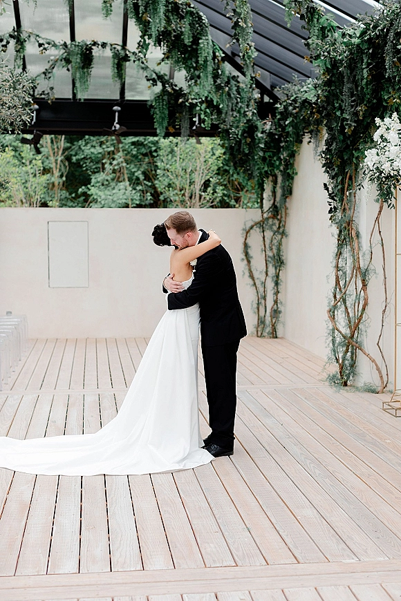 Wedding couple portrait of bride and groom hug beneath hanging greenery, her long train sweeping the wood deck in a bright greenhouse setting