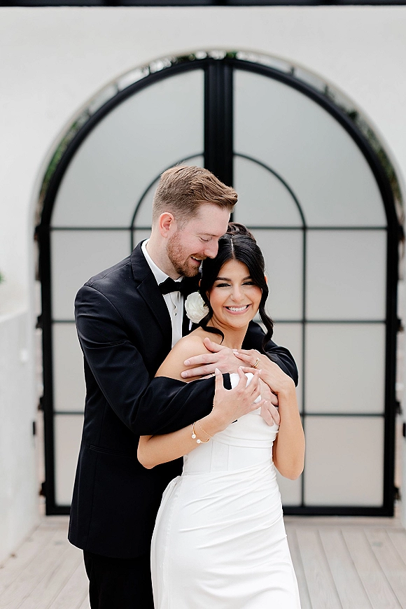 Couple portrait of bride and groom embrace, groom hugging bride in tuxedo and strapless dress before arched glass doors at venue patio