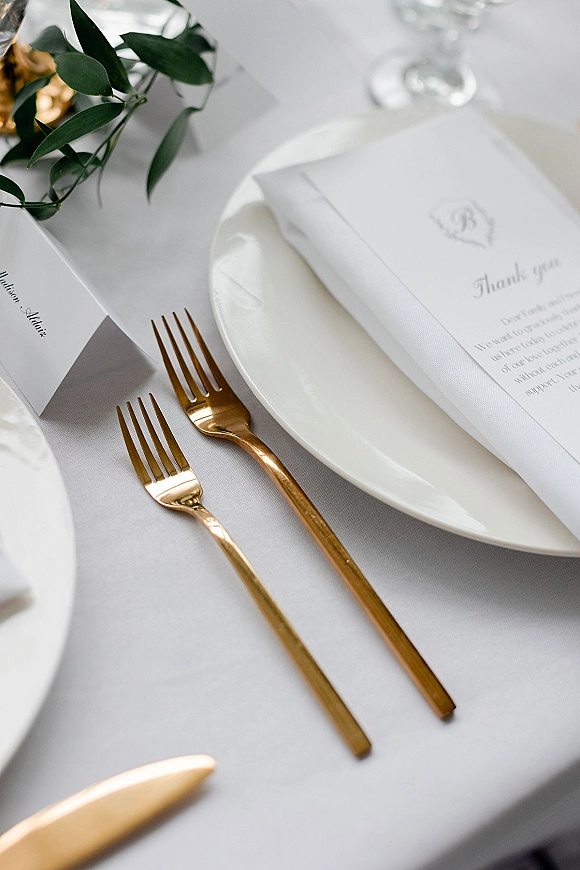 Wedding place setting with gold flatware, white plate, folded napkin, menu and place card on a white tablecloth with glassware and greenery