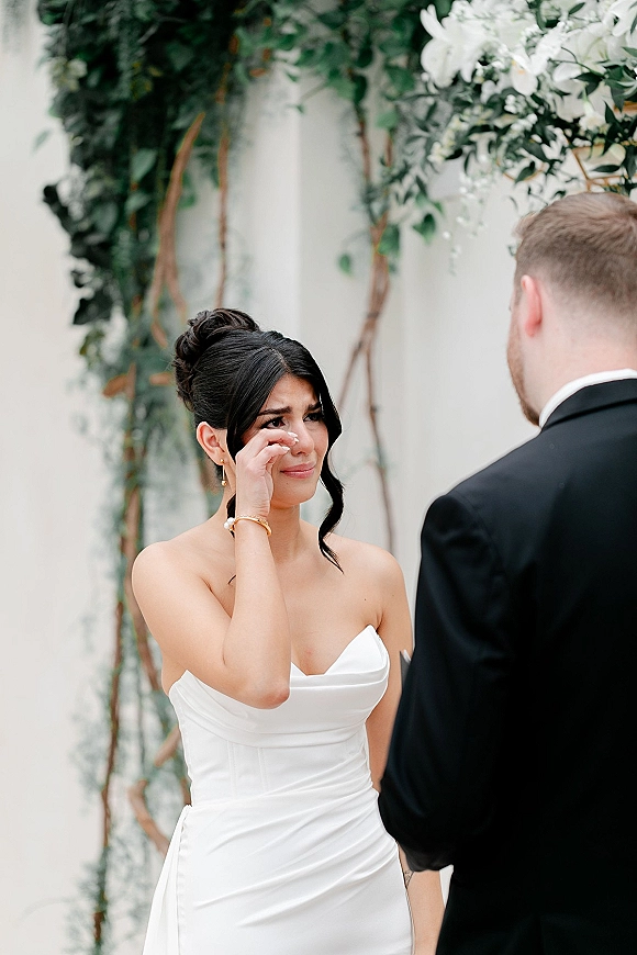 Wedding vows as bride wiping tears faces groom in tuxedo, wearing strapless dress and drop earrings against white wall with vines