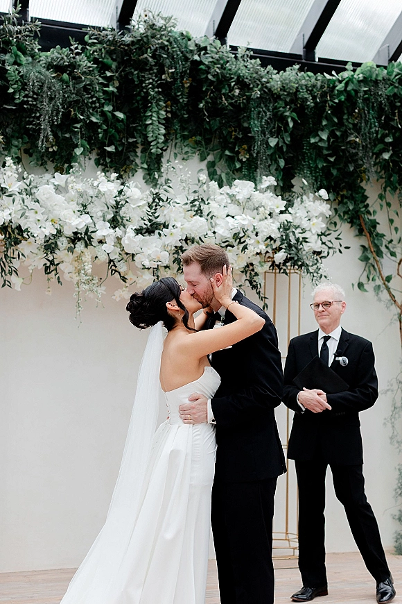 Wedding kiss as bride in a strapless dress and veil embraces groom in black tux beneath a greenery arch, officiant behind on white wall