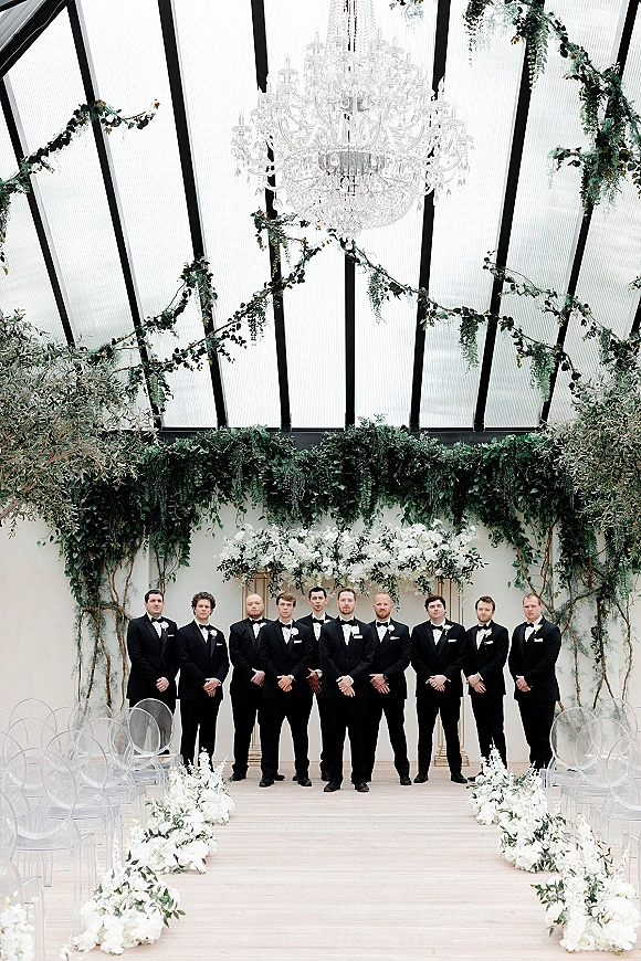 Groomsmen portrait of men in tuxedos lined up in black tuxedos with boutonnieres under a glass ceiling aisle with greenery garlands and chandelier