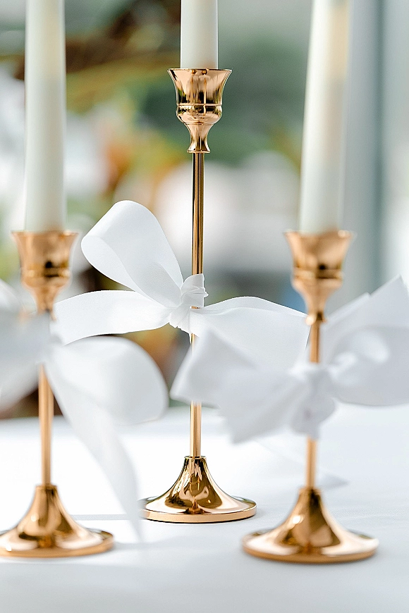 Wedding candle centerpiece with gold candlestick centerpiece, white taper candles and ribbon bows on a white tablecloth in soft window light