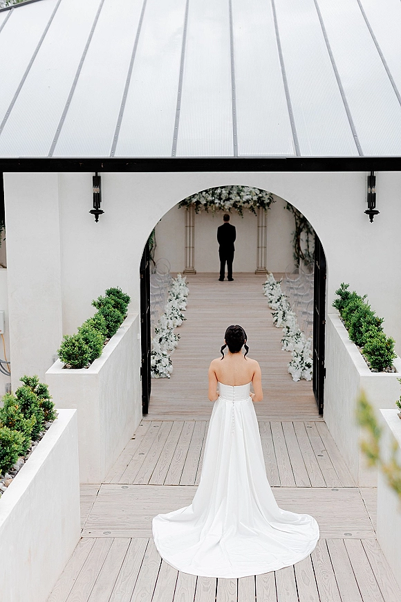 Wedding first look as bride walking to groom, her wedding dress train trailing down a wood walkway toward arch florals in a white stucco courtyard