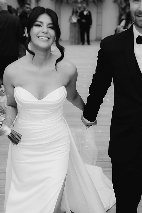 Couple portrait of bride and groom holding hands, smiling with bouquet and veil on an outdoor walkway as wedding guests watch behind