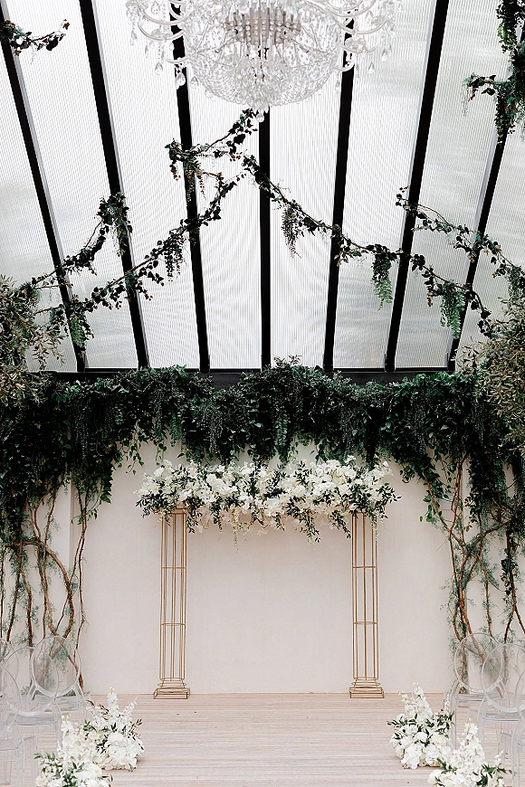 Ceremony backdrop with wedding altar flowers on gold frames, hanging greenery and chandelier under a glass ceiling with clear chairs