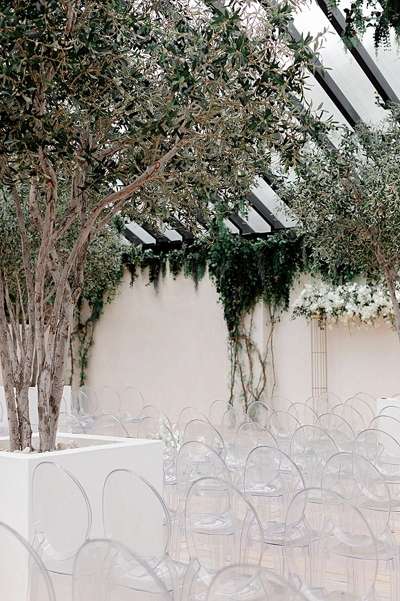 Ceremony seating with clear acrylic chairs lined beside white planters and olive trees under pergola beams against a white wall backdrop