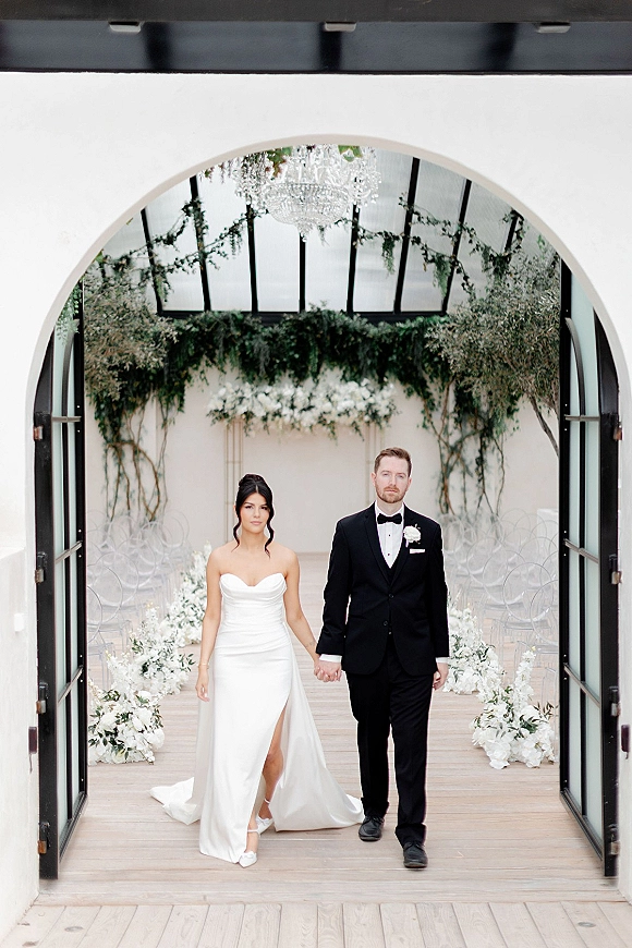 Couple portrait of bride and groom holding hands in a strapless satin gown and black tux, framed by chandelier and white aisle florals