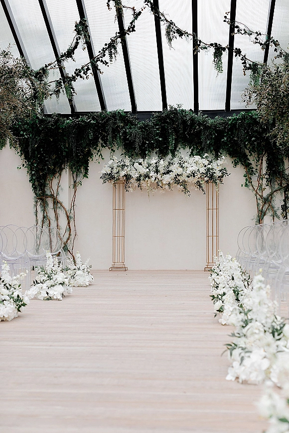 Ceremony setup with indoor wedding ceremony aisle, gold arch, white floral meadow and hanging greenery beneath a glass ceiling