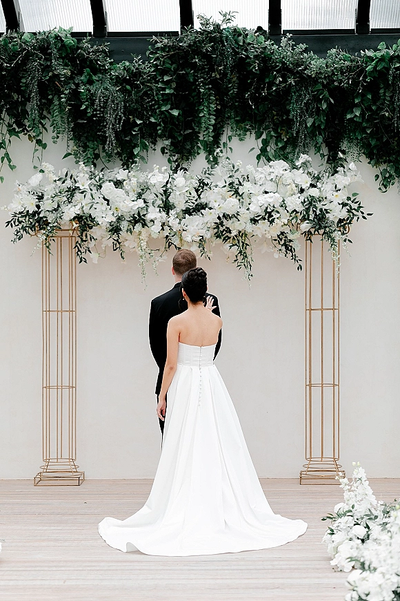 Ceremony moment as bride and groom at altar, facing officiant beneath white florals and greenery garland on gold columns indoors