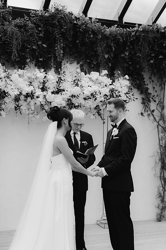 Wedding vows as bride and groom hold hands, veil and tuxedo with bow tie, before an officiant under a glass ceiling and greenery garland