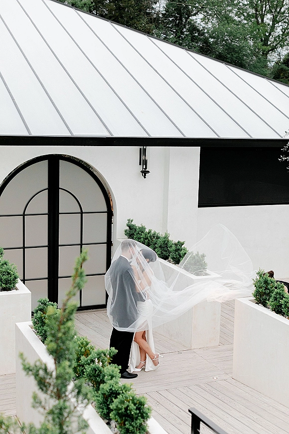 Wedding kiss beneath a billowing veil, bride in a short dress and heels embracing groom in a suit outside a white chapel with black arched doors